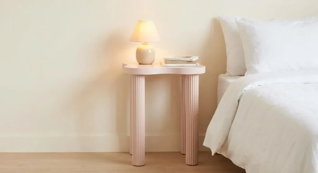  A slim, pale pink nightstand with a wave-shaped top and fluted legs, holding a small lamp and books next to a bed.